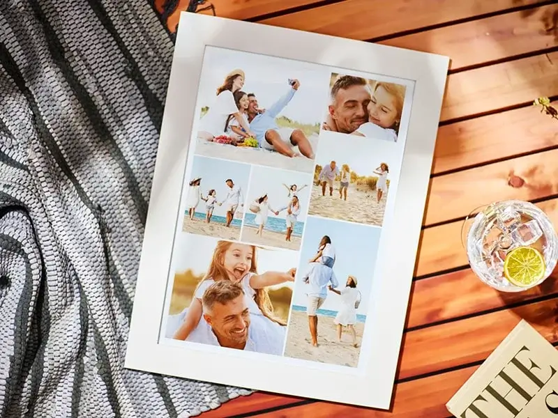 A framed collage of six photos showing people enjoying a day at the beach. The collage is placed on a wooden surface next to a striped blanket, a glass of water with lime slices, and part of a newspaper.