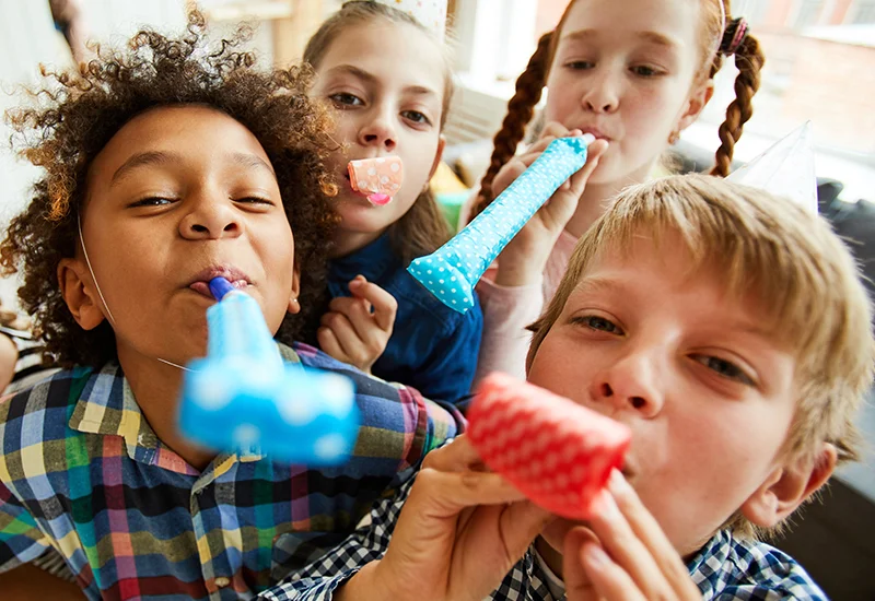 Four children blowing colourful party blowers towards the camera during a birthday celebration.