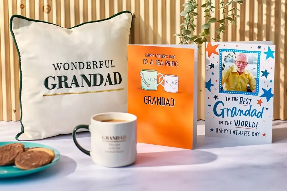 The image shows a cozy setup with a white pillow that has 'WONDERFUL GRANDAD' written on it, an orange Father's Day card with illustrations of tea cups and the text 'HAPPY FATHER'S DAY TO A TEA-RIFFIC GRANDAD,' another Father's Day card with stars and the text 'TO THE BEST Grandad IN THE WORLD! HAPPY FATHER'S DAY,' a white mug labeled 'GRANDAD,' and a plate with two cookies. The image shows a cozy setup with a white pillow that has 'WONDERFUL GRANDAD' written on it, an orange Father's Day card with illustrations of tea cups and the text 'HAPPY FATHER'S DAY TO A TEA-RIFFIC GRANDAD,' another Father's Day card with stars and the text 'TO THE BEST Grandad IN THE WORLD! HAPPY FATHER'S DAY,' a white mug labeled 'GRANDAD,' and a plate with two cookies.