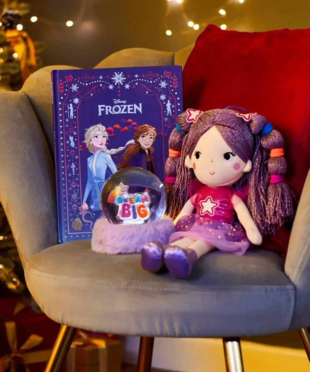 Purple Frozen-themed book, a plush doll with purple hair and pink dress, and a snow globe with the words Dream Big placed on a gray chair with a red cushion in a festive setting.