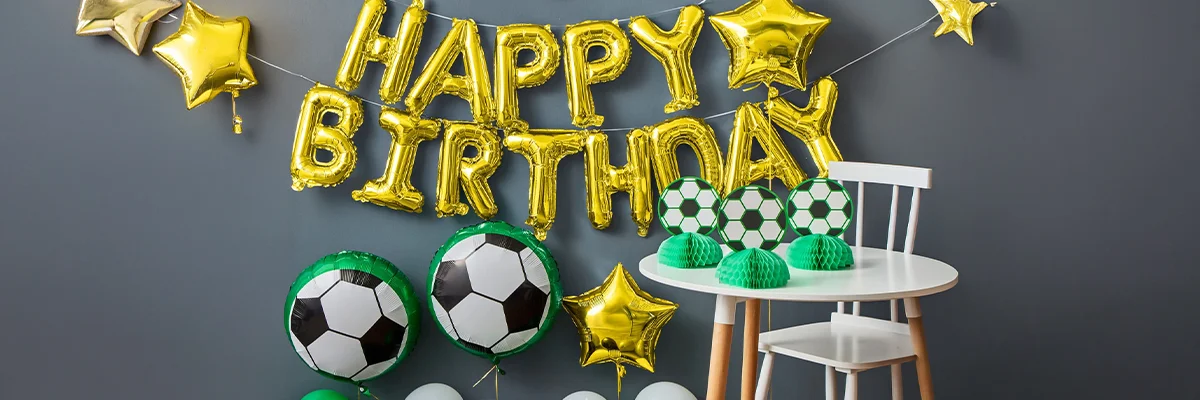 A football-themed birthday party setup featuring large gold foil letters spelling ‘Happy Birthday,’ gold star balloons, football foil balloons, green and white balloons, and football honeycomb centrepieces displayed on a white table against a dark grey wall. A football-themed birthday party setup featuring large gold foil letters spelling ‘Happy Birthday,’ gold star balloons, football foil balloons, green and white balloons, and football honeycomb centrepieces displayed on a white table against a dark grey wall.
