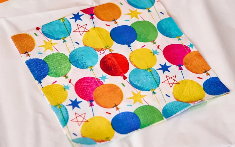 A folded paper party napkin featuring a colourful balloon pattern in blue, green, yellow, orange, pink and red, accented with stars and confetti motifs. The napkin is displayed on a white table covering and coordinates with matching birthday party tableware. A folded paper party napkin featuring a colourful balloon pattern in blue, green, yellow, orange, pink and red, accented with stars and confetti motifs. The napkin is displayed on a white table covering and coordinates with matching birthday party tableware.