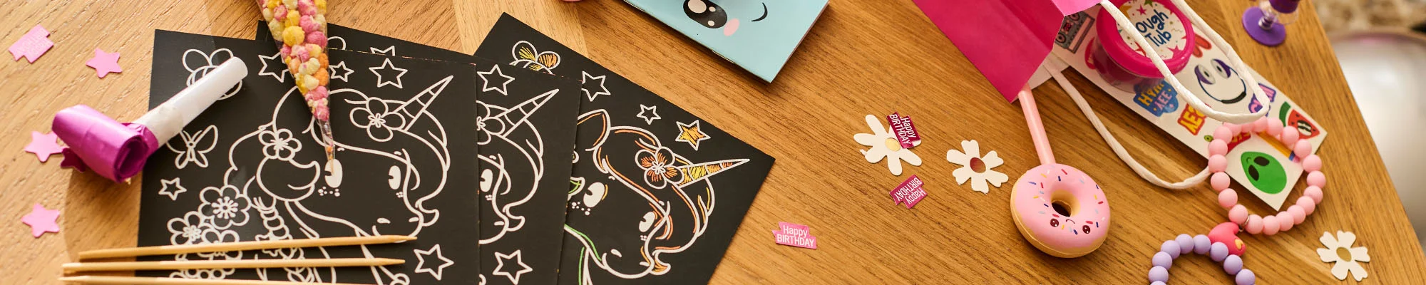 A close-up of princess-themed party bag fillers arranged on a wooden table, including unicorn scratch-art activity sheets with wooden styluses, a party blower, colourful sweet cones, novelty toys, bracelets, confetti shapes and a pink party favour bag. A close-up of princess-themed party bag fillers arranged on a wooden table, including unicorn scratch-art activity sheets with wooden styluses, a party blower, colourful sweet cones, novelty toys, bracelets, confetti shapes and a pink party favour bag.