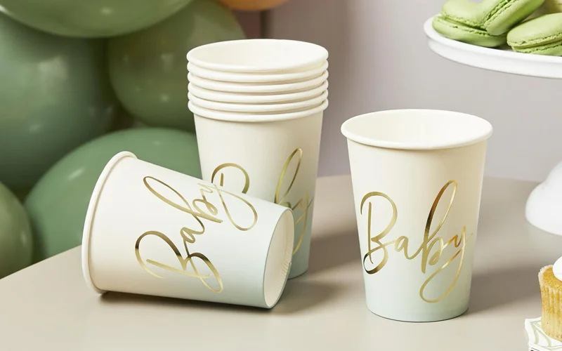 A stack of white paper cups featuring gold script lettering reading ‘Baby,’ displayed beside a sage green balloon garland and baby shower treats.