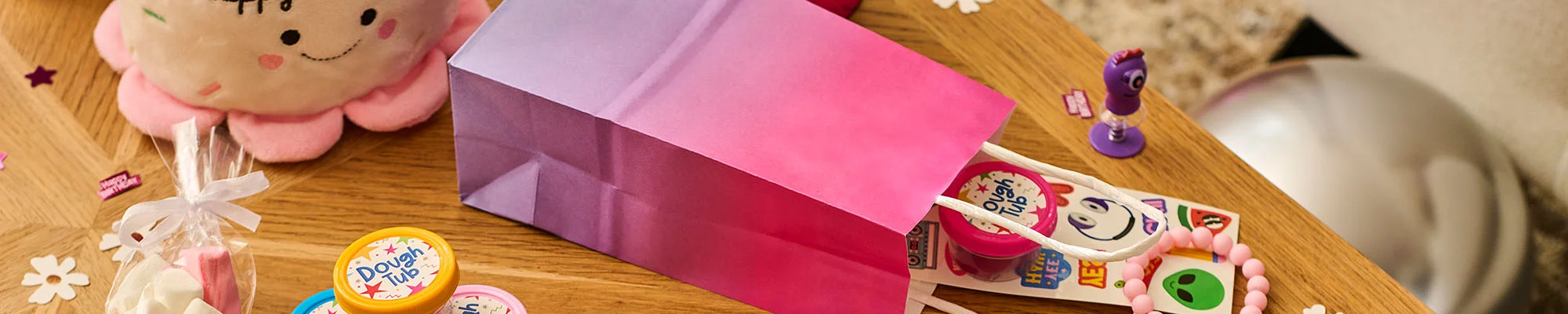 A pink party favour bag tipped on its side on a wooden table, revealing children's party bag fillers including a tub of dough, a sticker sheet with colourful characters and icons, novelty toys, and beaded bracelets. A cone of marshmallows, a flower-shaped plush cushion and birthday-themed confetti are scattered around the display. A pink party favour bag tipped on its side on a wooden table, revealing children's party bag fillers including a tub of dough, a sticker sheet with colourful characters and icons, novelty toys, and beaded bracelets. A cone of marshmallows, a flower-shaped plush cushion and birthday-themed confetti are scattered around the display.