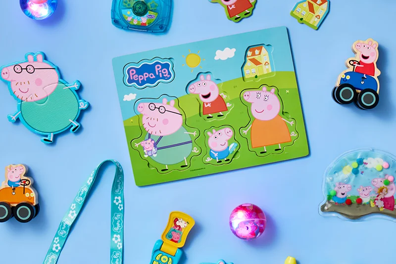 A Peppa Pig wooden jigsaw puzzle showing Peppa Pig family characters in an outdoor scene with grass, hills, houses and sunshine, surrounded by matching Peppa Pig toys and accessories arranged on a light blue background.