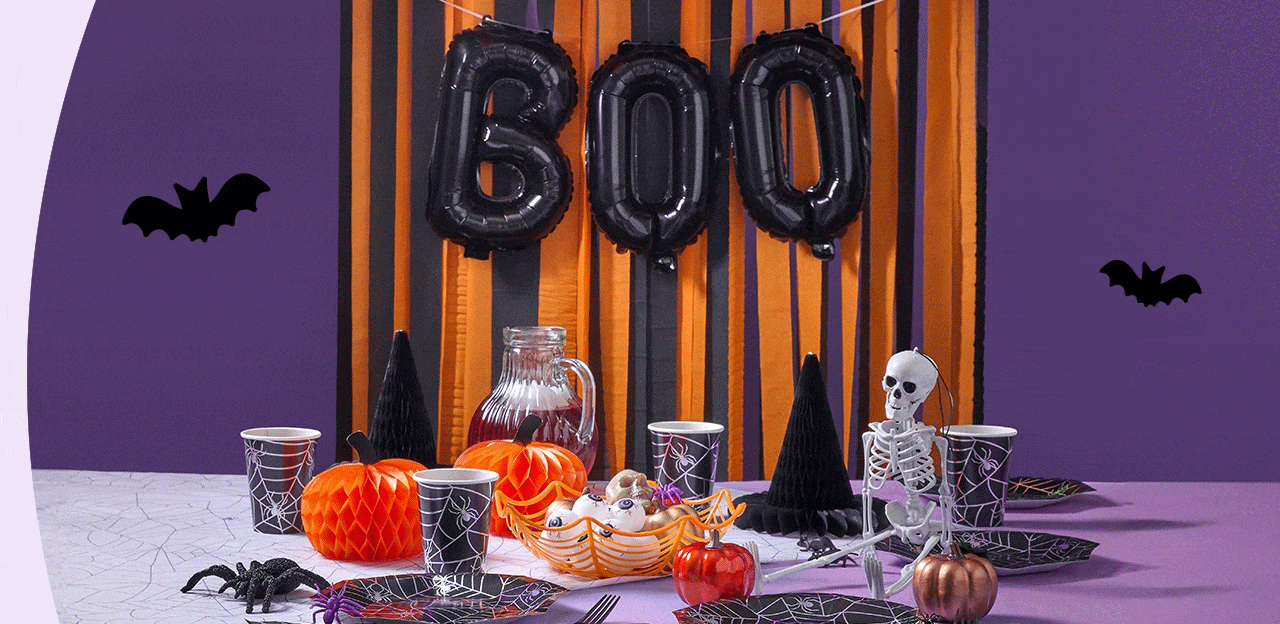 Halloween-themed setup with skulls, pumpkins, spider webs, a personalized mug and candle, candy bowl, and festive decorations against a purple backdrop.