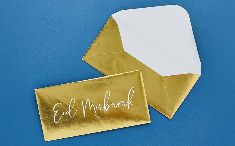 A gold foil envelope and matching card displayed on a blue background, with the card printed with the message ‘Eid Mubarak’ in white script.