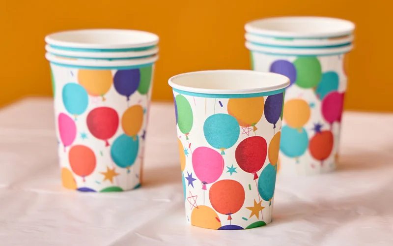 AA set of paper party cups decorated with colourful balloon illustrations in pink, blue, green, yellow, orange and red, accented with star and confetti details. The cups are displayed on a white table covering against a bright orange background as part of a coordinated birthday party tableware collection. A set of paper party cups decorated with colourful balloon illustrations in pink, blue, green, yellow, orange and red, accented with star and confetti details. The cups are displayed on a white table covering against a bright orange background as part of a coordinated birthday party tableware collection.