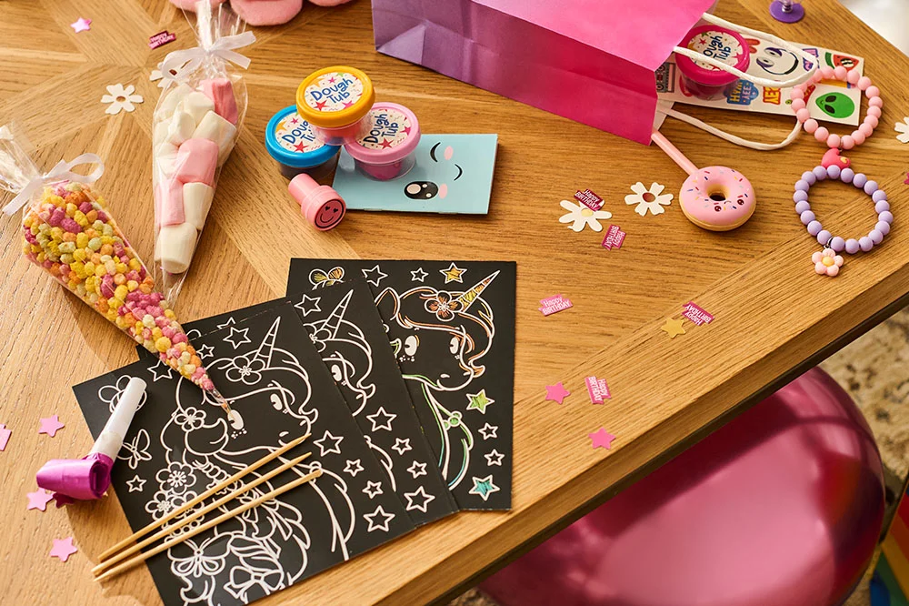 A selection of princess party bag fillers displayed on a wooden table, featuring unicorn scratch-art activity cards, sweet cones, tubs of modelling dough, novelty stationery, bracelets, small toys, confetti decorations and a pink party favour bag. A selection of princess party bag fillers displayed on a wooden table, featuring unicorn scratch-art activity cards, sweet cones, tubs of modelling dough, novelty stationery, bracelets, small toys, confetti decorations and a pink party favour bag.