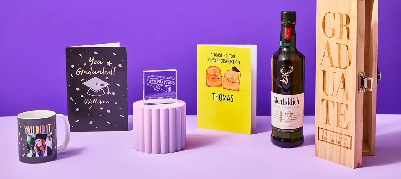 A graduation-themed display featuring a mug, greeting cards, a trophy, a bottle of Glenfiddich whiskey, and a wooden box with the word GRADUATE engraved on it.