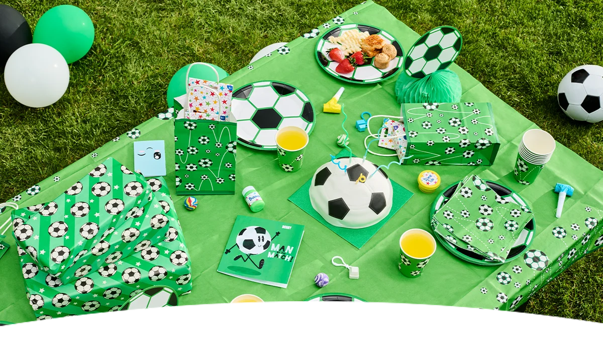 Add alt text here A football-themed party table set up on green grass, decorated with matching football-print plates, cups, napkins and gift bags. The display includes wrapped presents, balloons, a football-shaped cake, snacks, party favours, and a green tablecloth patterned with football designs.
