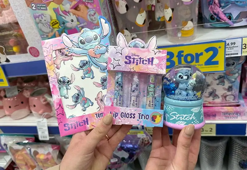 A selection of Disney Stitch themed gifts including a lip gloss trio, notebook, and a snow globe decoration held in hands in a shop display. A selection of Disney Stitch themed gifts including a lip gloss trio, notebook, and a snow globe decoration held in hands in a shop display.