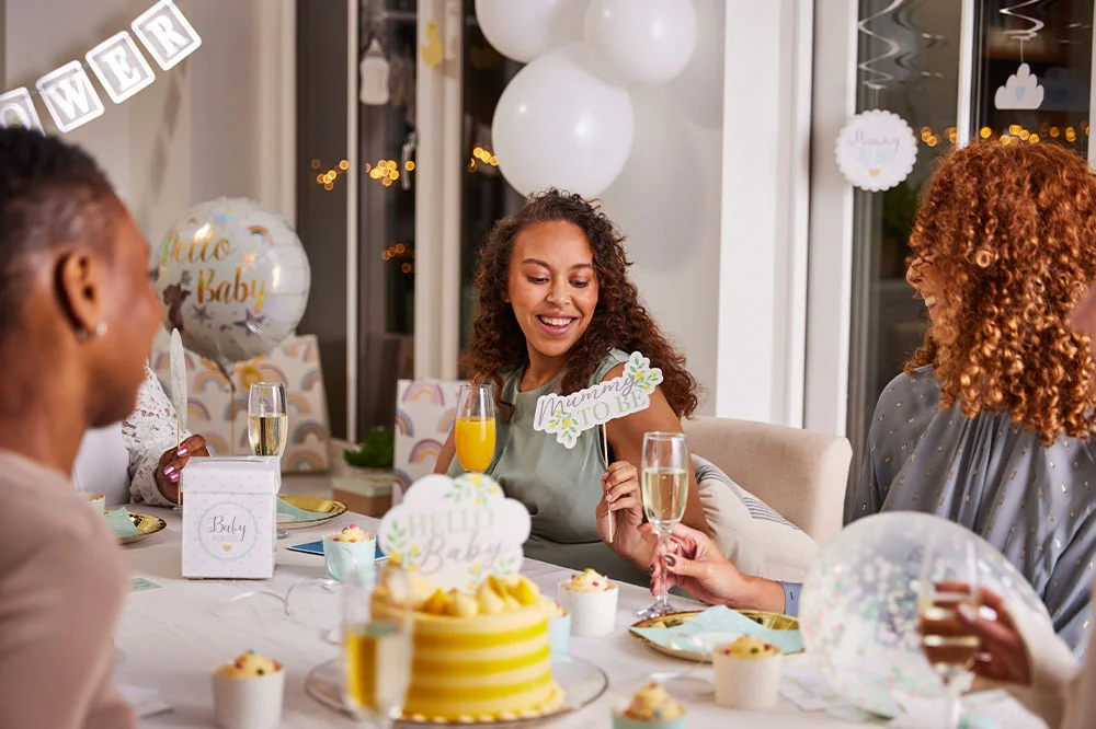 A baby shower gathering around a decorated table with cupcakes, drinks and baby-themed tableware. The display includes a ‘Mummy to Be’ sign, a ‘Hello Baby’ balloon, gift bags, white balloons and baby shower decorations, creating a celebration setup for the parent-to-be.
