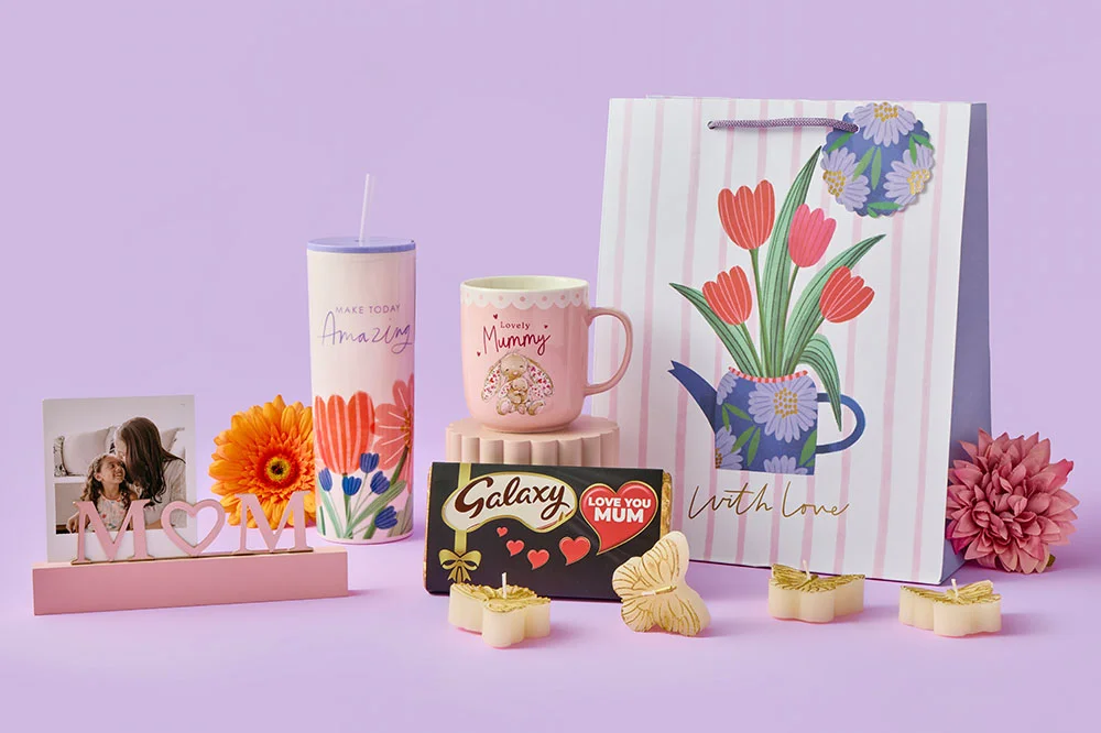 A selection of Mother’s Day gifts arranged on a pastel purple background, including a floral travel tumbler, a pink mug, a large striped gift bag with tulip artwork, a chocolate bar, artificial flowers, and small decorative keepsakes.