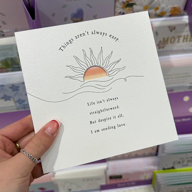 A square greeting card held in one hand, featuring a line‑art sun rising over waves with the message: ‘Things aren’t always easy. Life isn’t always straightforward. But despite it all, I am sending love.