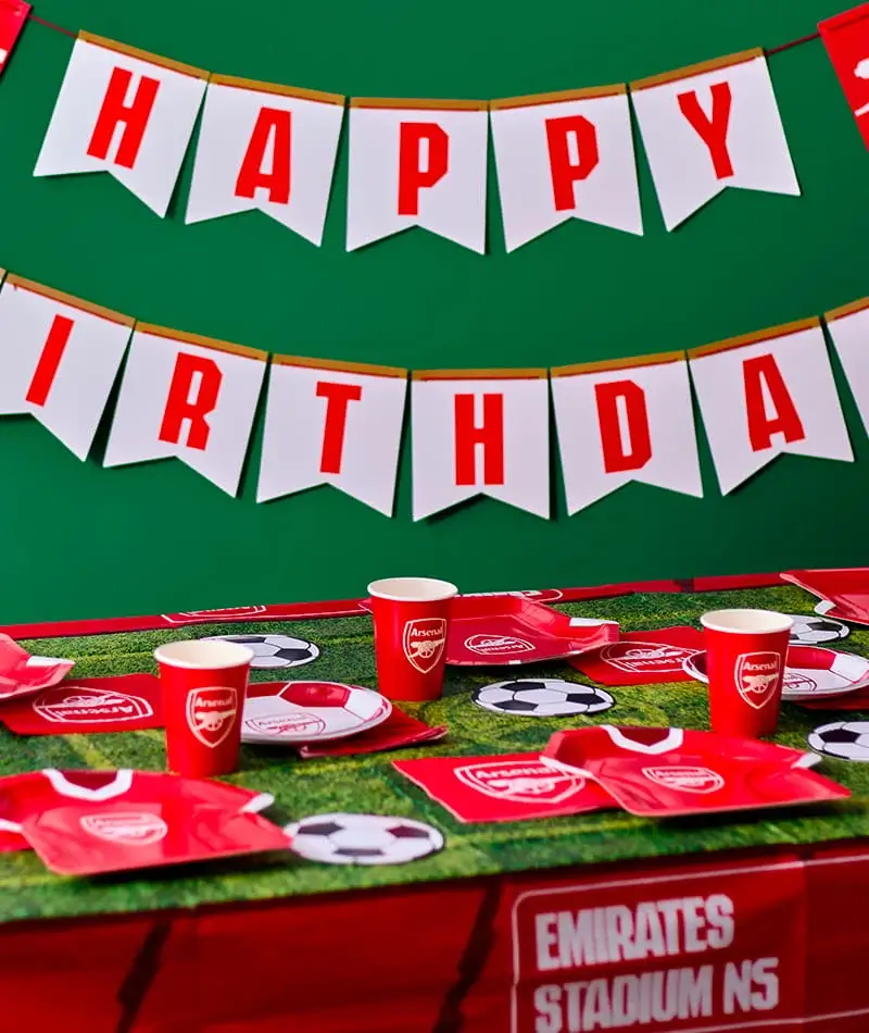 An Arsenal football-themed birthday party table display featuring red branded plates, cups and napkins arranged on a grass-effect table covering. A ‘Happy Birthday’ bunting banner hangs against a green background, with football-shaped balloons at either side and football-themed table decorations completing the celebration setup. An Arsenal football-themed birthday party table display featuring red branded plates, cups and napkins arranged on a grass-effect table covering. A ‘Happy Birthday’ bunting banner hangs against a green background, with football-shaped balloons at either side and football-themed table decorations completing the celebration setup.