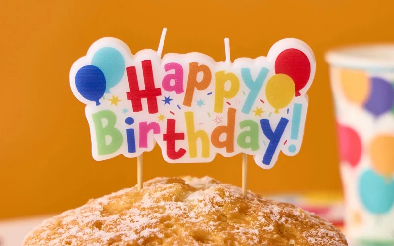 A colourful ‘Happy Birthday!’ cake topper decorated with balloon illustrations, displayed on top of a sugar-dusted cake against a bright orange background.