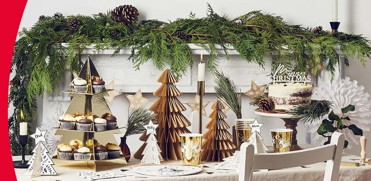 Festive Christmas table setting with paper trees, candles, greenery, cupcakes, a cake with Merry Christmas topper, personalized tree-shaped place cards, and a decorated mantel with pinecones and evergreen branches. Festive Christmas table setting with paper trees, candles, greenery, cupcakes, a cake with Merry Christmas topper, personalized tree-shaped place cards, and a decorated mantel with pinecones and evergreen branches.