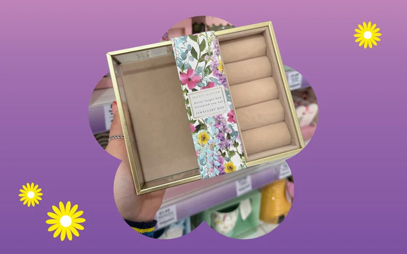 A rectangular jewellery box with a clear lid and beige interior, wrapped with a floral patterned band, held up in a store aisle against a purple and pink gradient background.