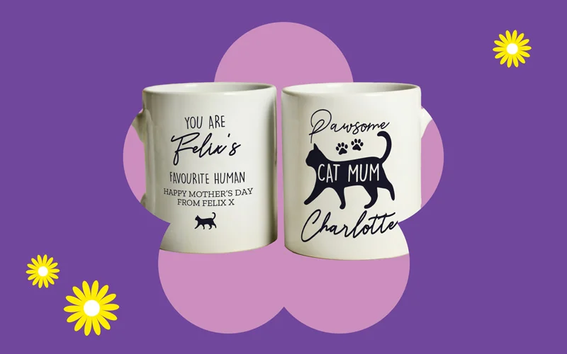 Two white mugs with cat‑themed designs and messages, including ‘You are Felix’s favourite human’ and ‘Pawsome Cat Mum Charlotte,’ displayed against a purple background with a pink flower shape and yellow daisies.