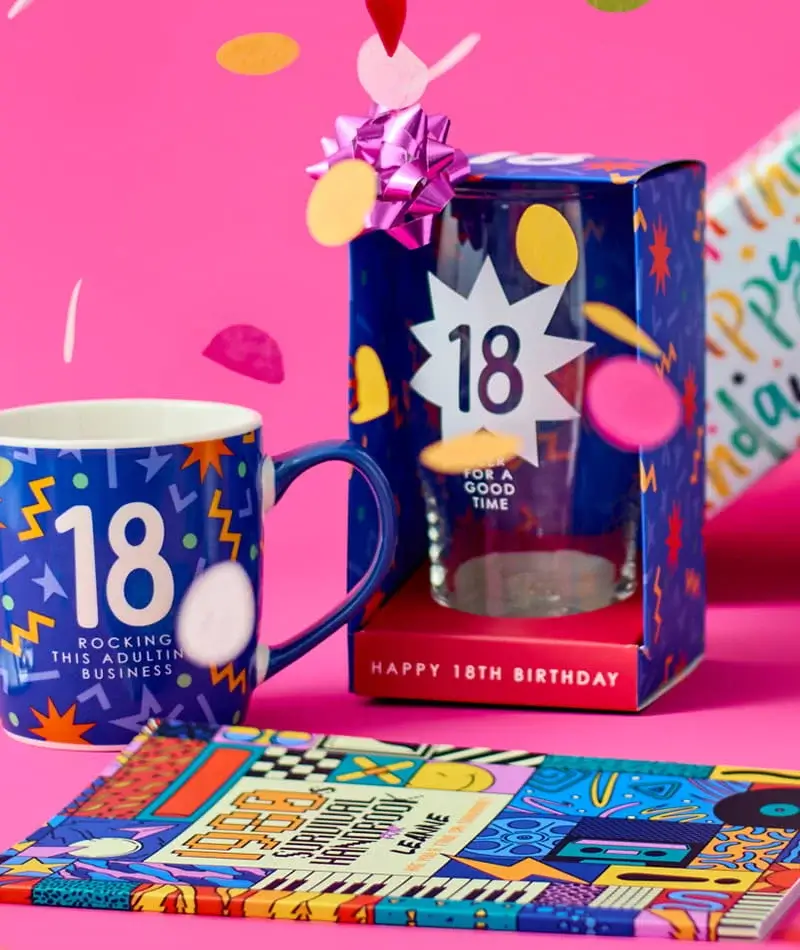 Colourful 18th birthday gift set including a blue mug, boxed pint glass, notebook, and confetti on a pink background