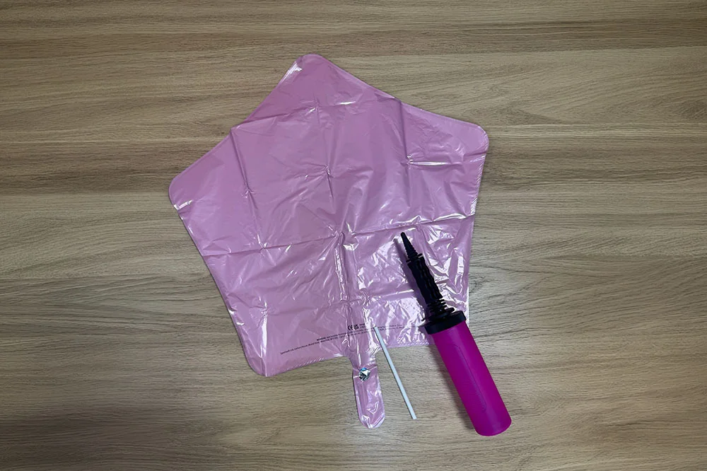 A close-up of a pink star-shaped foil balloon before inflation, displayed on a wooden surface alongside a pink hand pump and white balloon stick. The image shows the equipment used to inflate and assemble foil balloon decorations for party displays. A close-up of a pink star-shaped foil balloon before inflation, displayed on a wooden surface alongside a pink hand pump and white balloon stick. The image shows the equipment used to inflate and assemble foil balloon decorations for party displays.