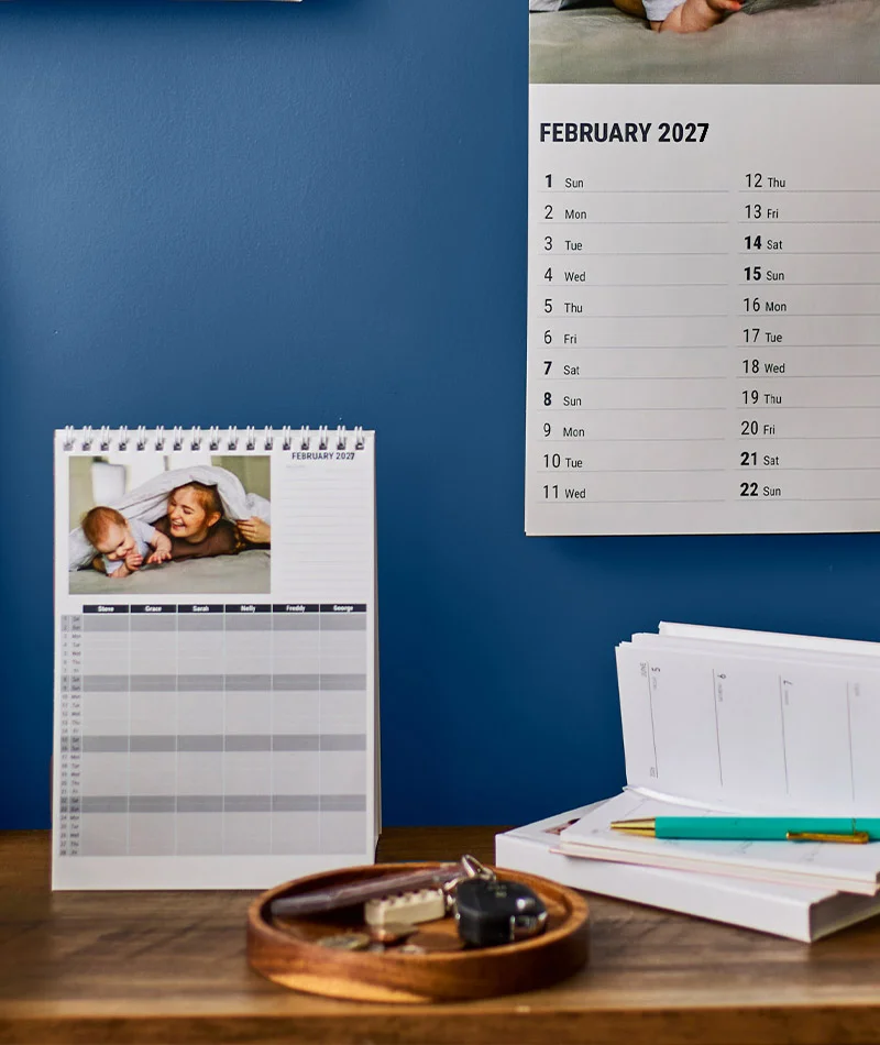 Desk stationery collection featuring a personalised wall calendar, notebooks, weekly planner, diffuser, and mug arranged on a wooden desk.