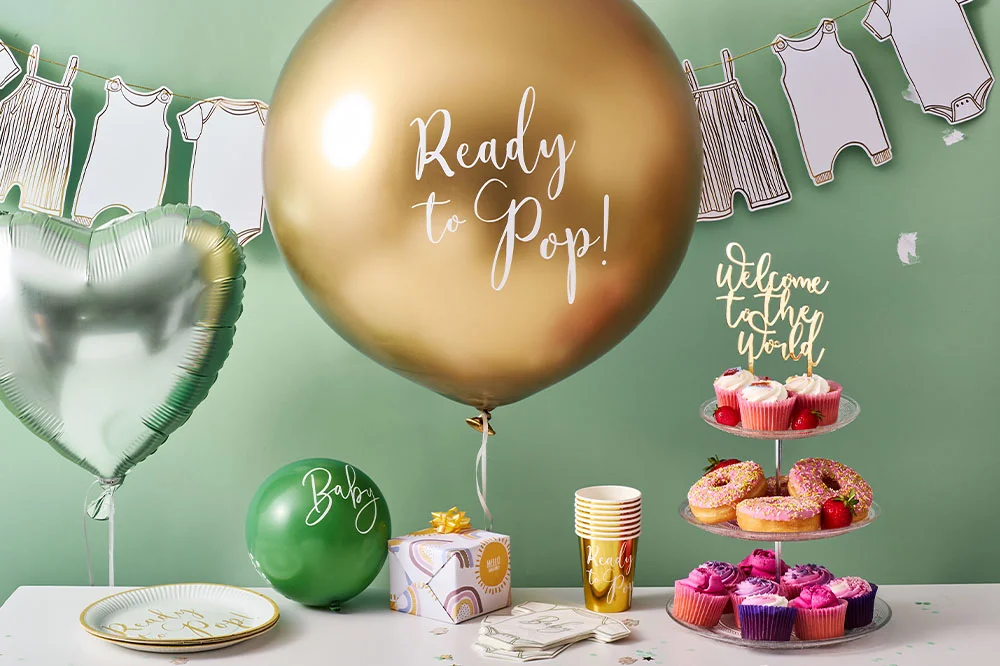 A baby shower dessert table decorated with a giant gold ‘Ready to Pop!’ balloon, a silver heart-shaped foil balloon and baby-themed tableware. The display includes cupcakes, pink doughnuts, personalised cups, a gift box, baby-clothes bunting and a ‘Welcome to the World’ sign against a sage green background.