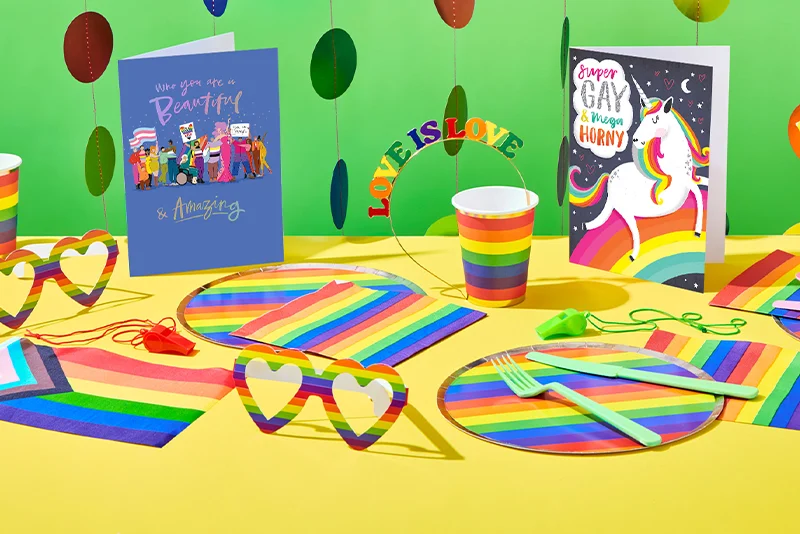 A wide Pride‑themed display arranged on a yellow tabletop with a green backdrop, featuring rainbow party decorations and stationery including striped plates, rainbow cups, colourful paper straws, whistles, and heart‑shaped sunglasses, alongside two greeting cards reading ‘Who you are is Beautiful & Amazing’ and ‘Super Gay & Mega Horny’ illustrated with a unicorn, with a rainbow ‘Love is Love’ sign in the centre.