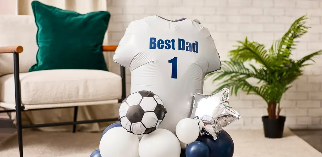 'A white balloon shaped like a soccer jersey with the text 'Best Dad' and the number '1' printed on it. The balloon is surrounded by other balloons, including a soccer ball-shaped balloon, white and blue round balloons, and a silver star-shaped balloon. In the background, there is a green cushion on a chair, a potted plant, and part of a brick wall.'