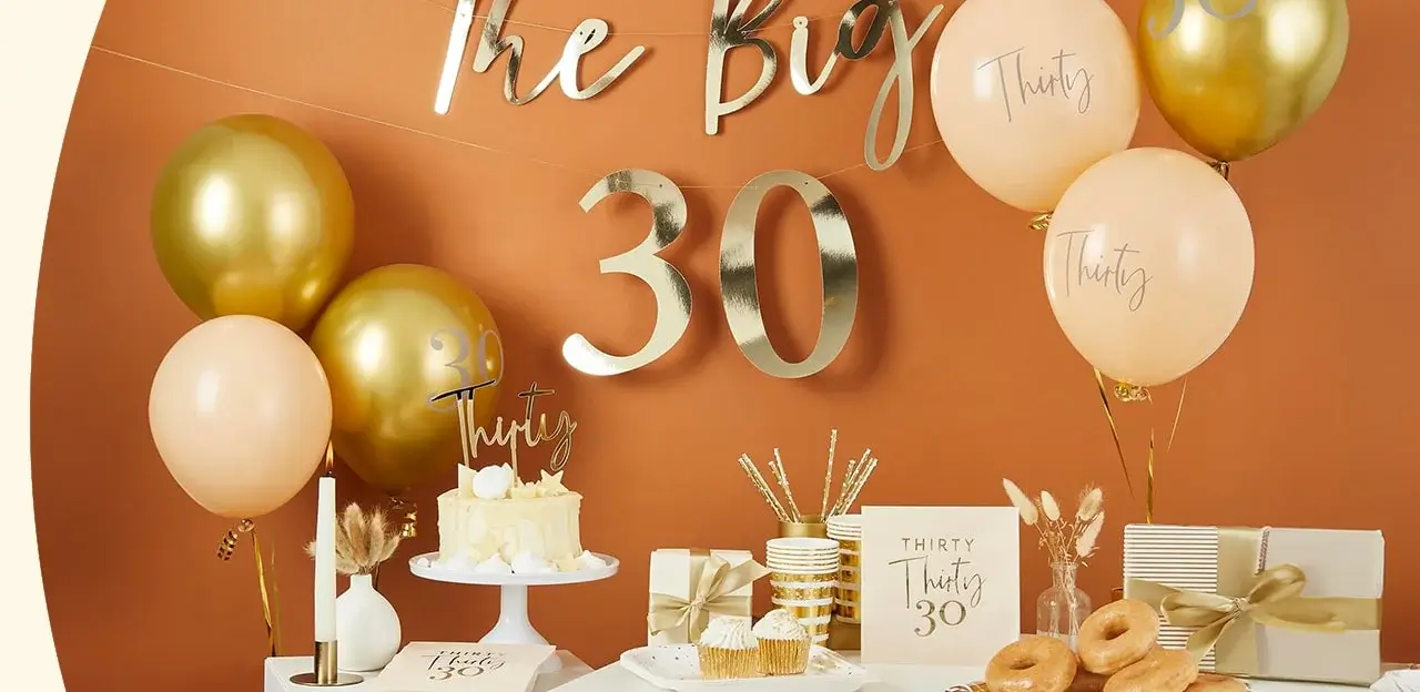 30th birthday celebration setup with gold and peach decorations, including balloons with 'Thirty', a cake, cupcakes, donuts, wrapped gifts, and a sign reading 'The Big 30' against an orange wall.