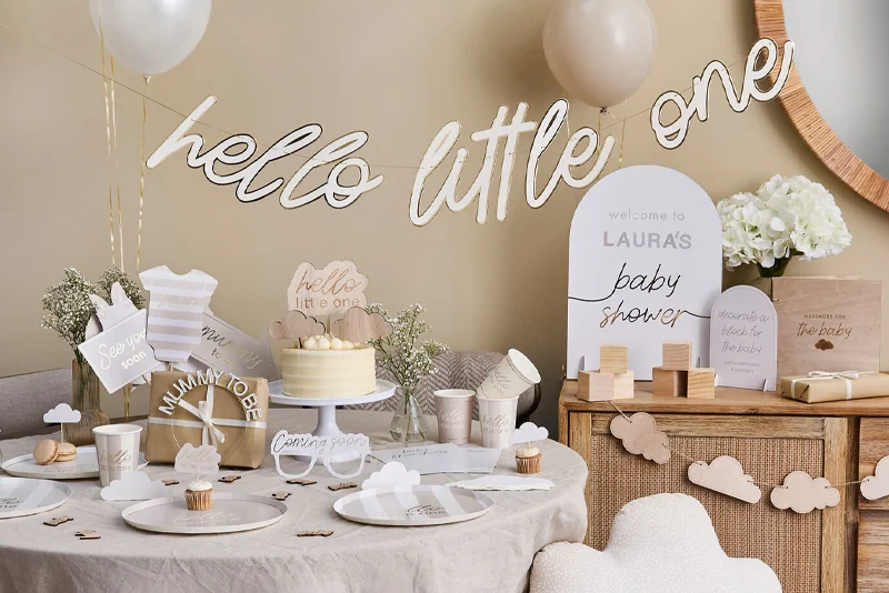 A neutral-toned baby shower setup featuring a ‘hello little one’ banner, personalised signage reading ‘Welcome to Laura’s baby shower,’ baby-themed tableware, cupcakes, balloons and decorative keepsakes. Wooden cloud decorations, soft neutral colours and baby shower games create a coordinated celebration display.