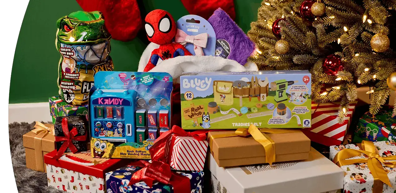 Selection of Christmas gifts and toys, including a Bluey playset, Spider-Man plush, novelty sweets and a miniature vending machine, displayed among wrapped presents beside a decorated Christmas tree.