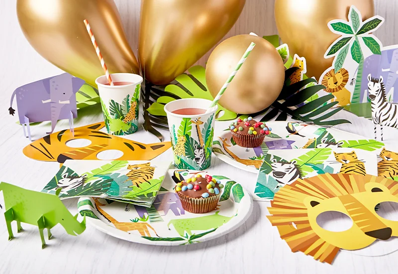 A safari-themed party table featuring jungle-print cups, plates and napkins, gold balloons, animal decorations, lion masks and colourful cupcakes.