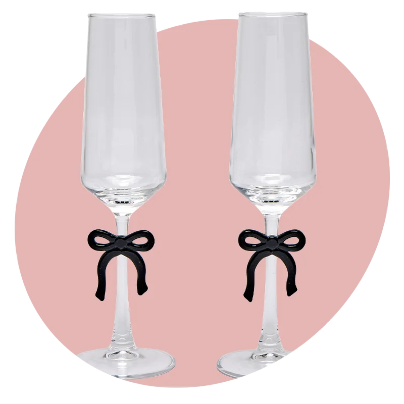 A pair of tall champagne flutes with clear glass and black bow details around the stems, displayed against a soft pink circular background. A pair of tall champagne flutes with clear glass and black bow details around the stems, displayed against a soft pink circular background.