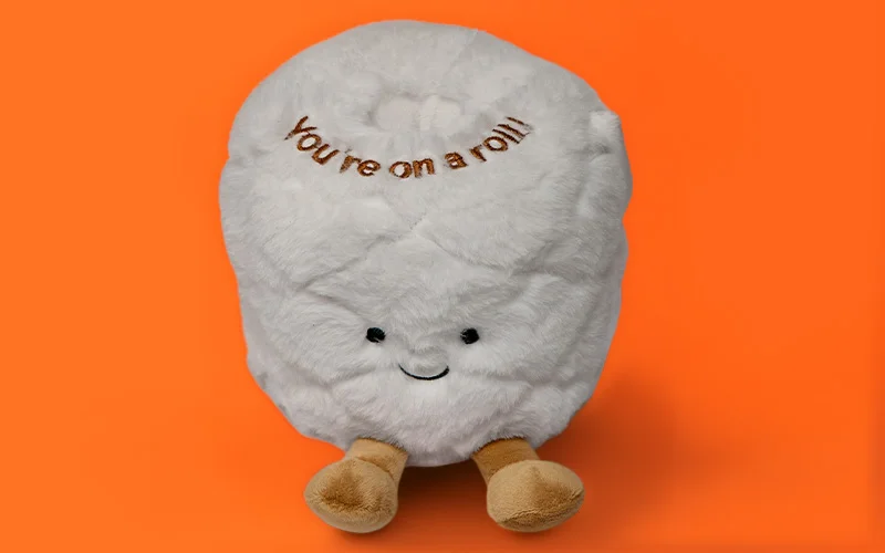 A soft novelty plush toy shaped like a white toilet roll with small embroidered features and the text ‘You’re on a roll,’ displayed against a bright orange background. A soft novelty plush toy shaped like a white toilet roll with small embroidered features and the text ‘You’re on a roll,’ displayed against a bright orange background.
