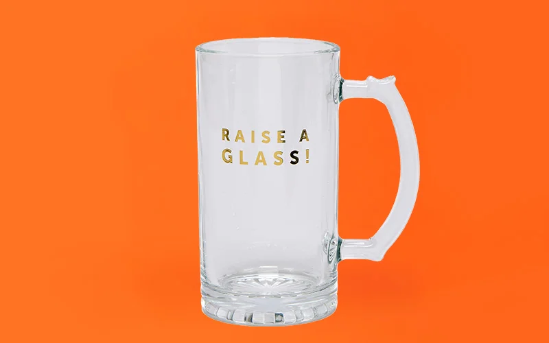 A clear glass beer mug with a sturdy handle and thick base, featuring gold lettering that reads ‘Raise a Glass!’ on the front, displayed against a bright orange background. A clear glass beer mug with a sturdy handle and thick base, featuring gold lettering that reads ‘Raise a Glass!’ on the front, displayed against a bright orange background.