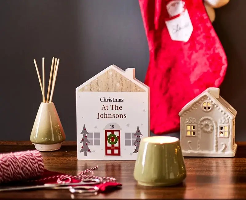 Festive Christmas scene with a decorated tree, gold ornaments, a red stocking, a lit candle, and ceramic house decorations on a wooden table.