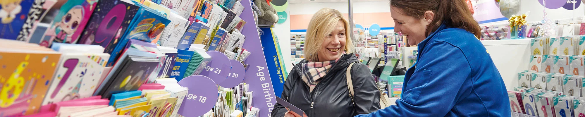 A wide view of a greeting card aisle showing two people looking at a card together, surrounded by shelves stocked with colourful cards and stationery.