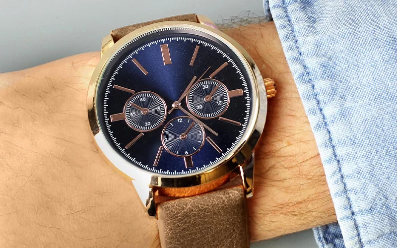 A close‑up of a wrist wearing a gold‑trimmed watch with a dark blue face, three sub‑dials, and a brown leather strap.