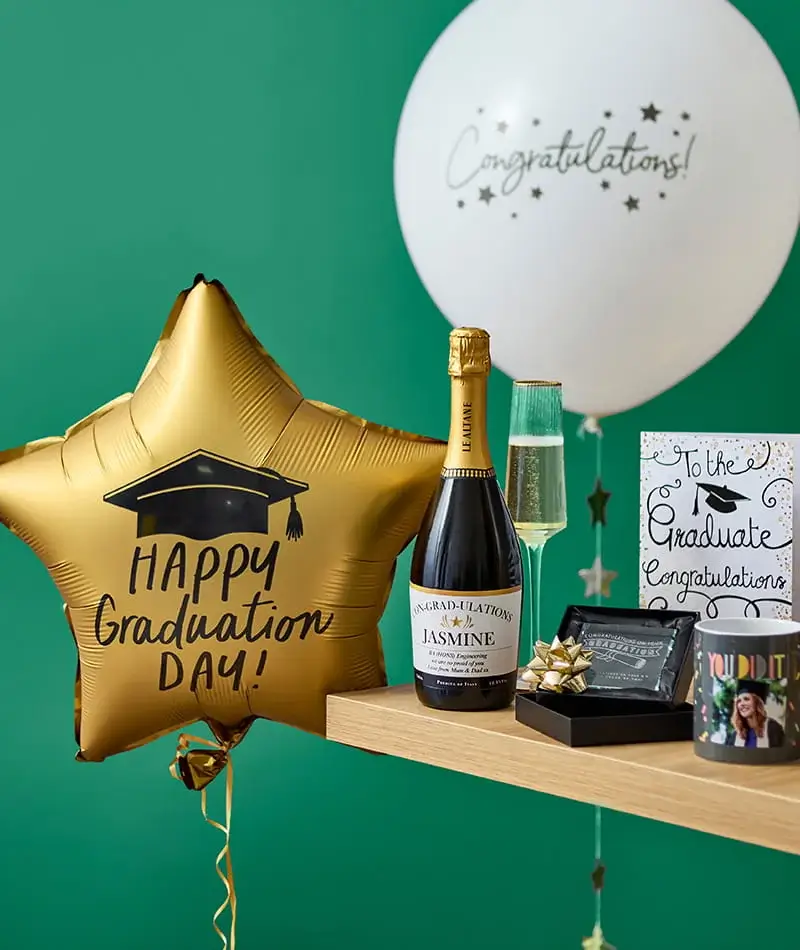Graduation celebration setup with gold graduation balloon, congratulations balloon, cards, champagne, and gift items arranged on a shelf