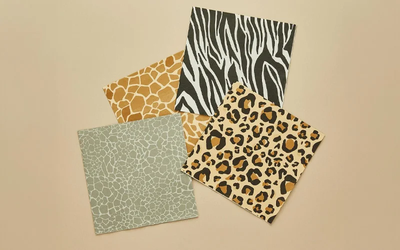 A set of safari-themed paper napkins displayed on a neutral beige background, featuring four different animal-inspired prints: zebra stripes, leopard spots, giraffe markings and a grey textured pattern. The napkins are arranged overlapping to showcase the coordinated safari party design. A set of safari-themed paper napkins displayed on a neutral beige background, featuring four different animal-inspired prints: zebra stripes, leopard spots, giraffe markings and a grey textured pattern. The napkins are arranged overlapping to showcase the coordinated safari party design.