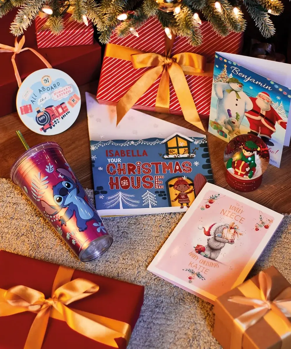Christmas-themed setup with a personalized pillow reading 'N is for NOAH', a red stocking labeled 'Louise', a plush bunny with 'My First Christmas' on its ears, wooden maracas, and an elf snow globe beside a decorated tree.