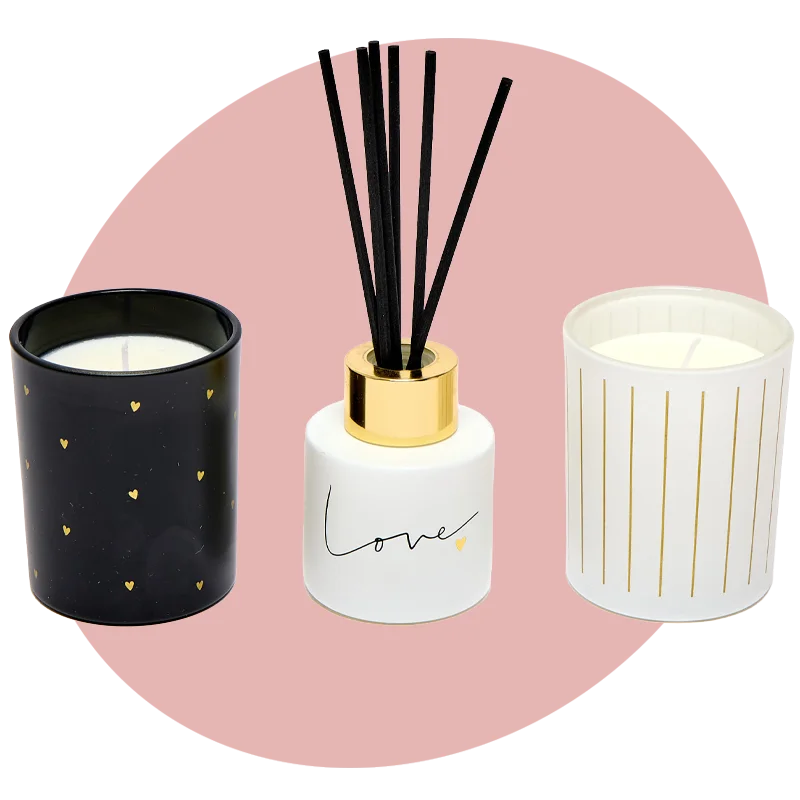 A home fragrance set including a black candle with gold hearts, a white reed diffuser with black reeds, and a white candle with gold stripe detailing, displayed against a soft pink circular background. A home fragrance set including a black candle with gold hearts, a white reed diffuser with black reeds, and a white candle with gold stripe detailing, displayed against a soft pink circular background.