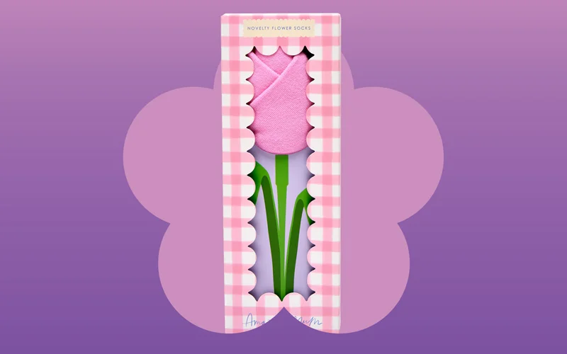 A boxed gift set containing novelty flower-shaped pink socks presented like a tulip, in pink gingham packaging, shown against a purple and pink gradient background with a flower silhouette.