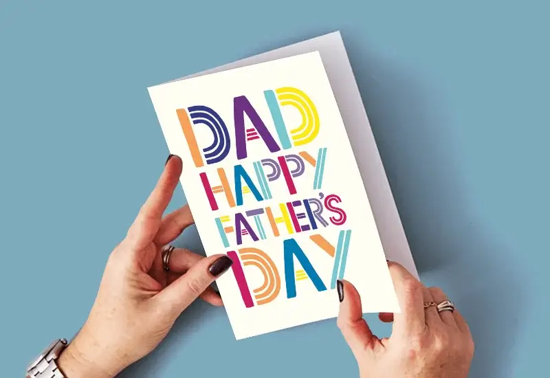 A person holding a Father's Day card with bold colorful letters saying DAD HAPPY FATHER'S DAY on a blue background.