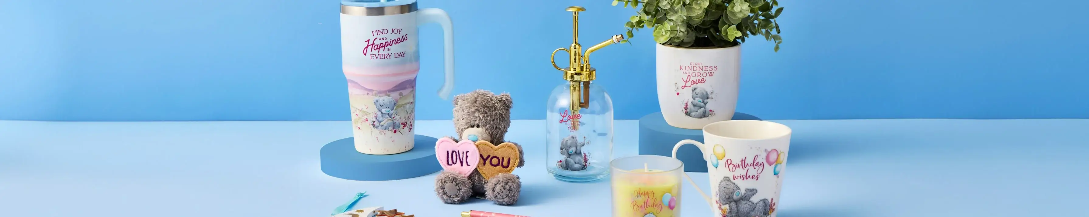 A collection of items on a light blue surface against a blue background. The items include a travel mug with the text 'FIND JOY Happiness EVERY DAY', a small teddy bear holding two hearts with the text 'LOVE YOU', a glass spray bottle with the text 'Love', a potted plant in a pot with the text 'KINDNESS GROWS Love', and a ceramic mug with the text 'Birthday wishes'. There is also an open notebook, some colored pens, and pencils.