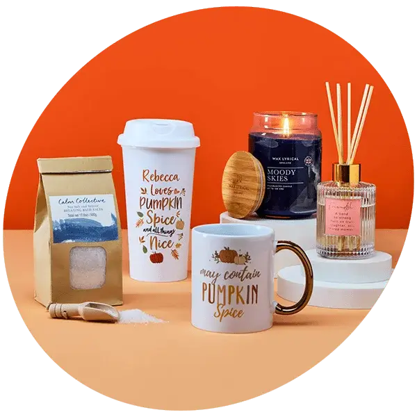 Autumn-themed gift set featuring pumpkin spice mugs, bath tea, a body brush, an orange candle, and a reed diffuser against an orange background.