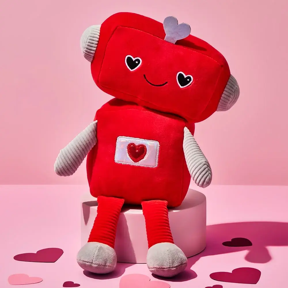 Red plush robot toy with heart-shaped eyes, a small heart antenna, and a stitched heart on its chest, sitting on a white round platform surrounded by pink and red paper hearts against a pink background.